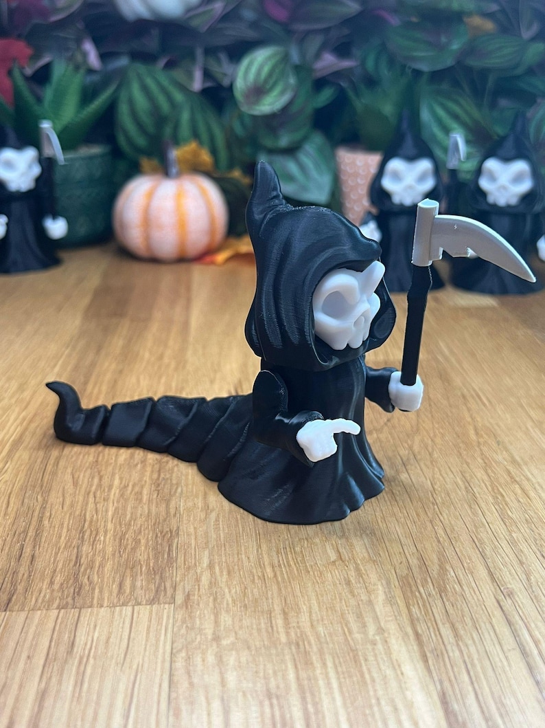 May include: A black and white 3D printed figurine of a skeletal figure in a hooded robe, holding a scythe. The figure is in a crawling pose with a long, segmented tail.