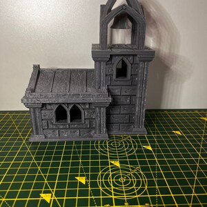 May include: A gray 3D printed miniature church with a bell tower and a small window. The church is sitting on a green cutting mat.