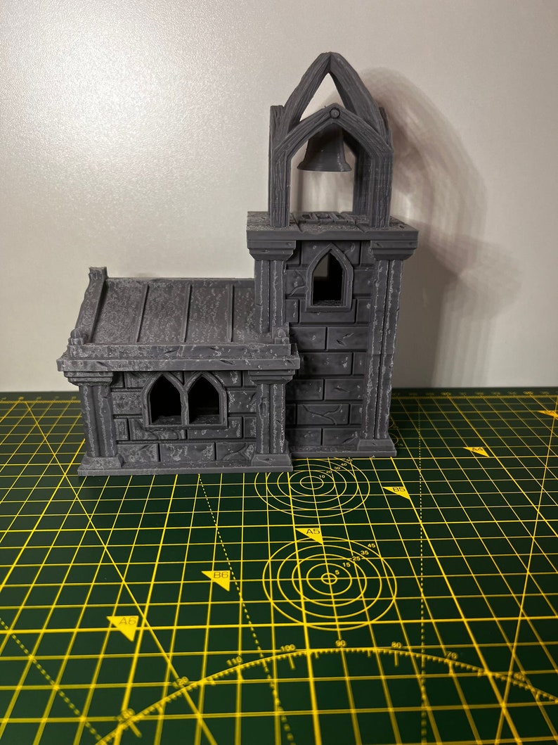 May include: A gray 3D printed miniature church with a bell tower and a small window. The church is sitting on a green cutting mat.