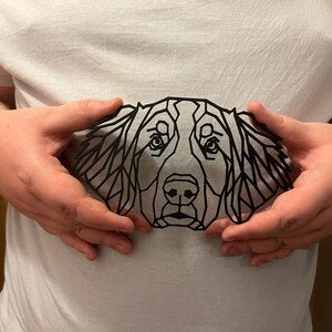 May include: A black geometric silhouette of a dog's head, with a detailed design of triangles and lines. The silhouette is held in front of a white shirt.