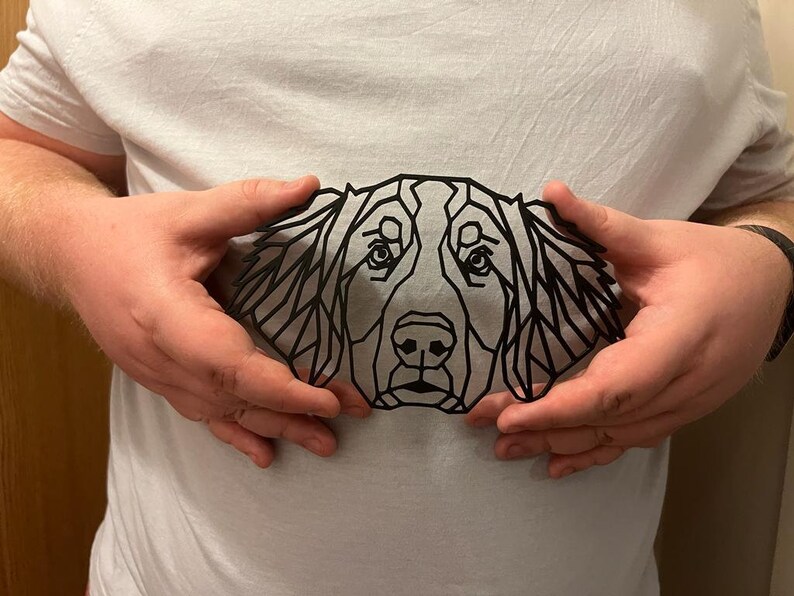 May include: A black geometric silhouette of a dog's head, with a detailed design of triangles and lines. The silhouette is held in front of a white shirt.