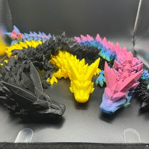 May include: Four 3D printed dragon figurines in black, yellow, pink, and blue. The dragons are posed in a variety of positions and have a detailed, textured surface.