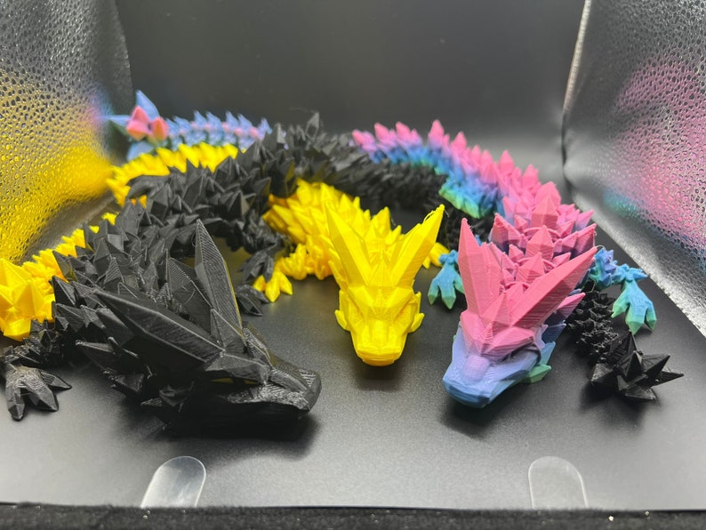 May include: Four 3D printed dragon figurines in black, yellow, pink, and blue. The dragons are posed in a variety of positions and have a detailed, textured surface.