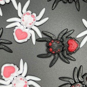 May include: A collection of eight 3D printed spiders, four black and four white. Each spider has a red heart on its abdomen and red dots on its body. The spiders are arranged in a pattern on a dark gray surface.