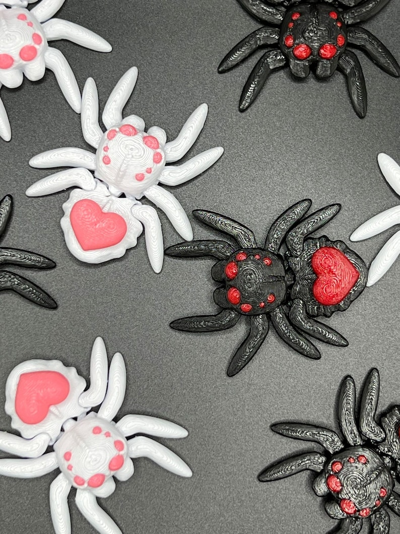 May include: A collection of eight 3D printed spiders, four black and four white. Each spider has a red heart on its abdomen and red dots on its body. The spiders are arranged in a pattern on a dark gray surface.