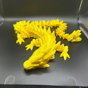 May include: A bright yellow 3D printed dragon figurine with a spiky, textured design. The dragon is posed in a curled position with its head resting on the ground.