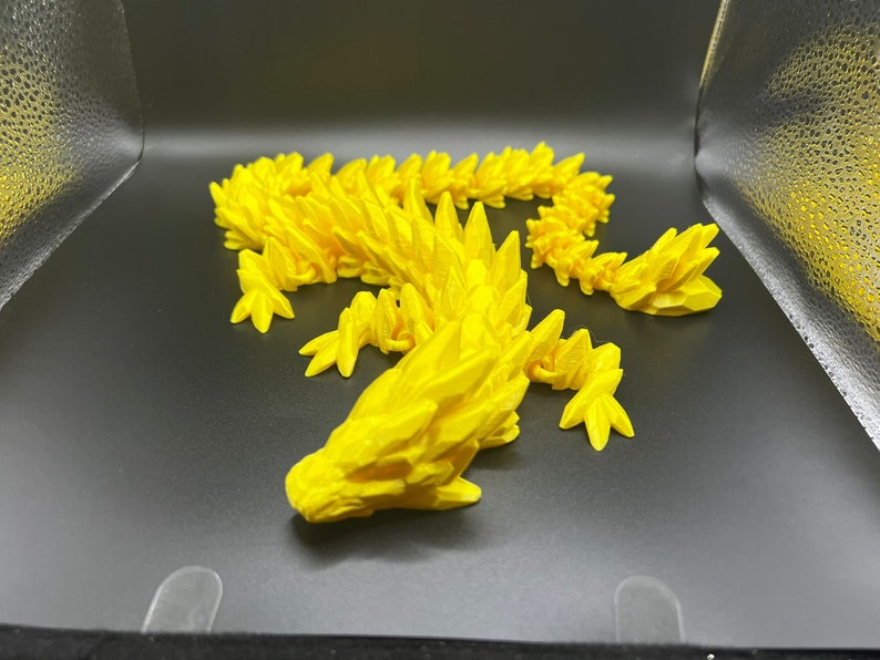 May include: A bright yellow 3D printed dragon figurine with a spiky, textured design. The dragon is posed in a curled position with its head resting on the ground.