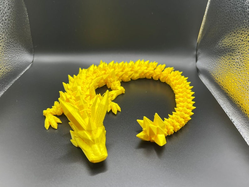 May include: A yellow 3D printed dragon figurine with a spiky, textured design. The dragon is curled into a circle with its tail touching its head.