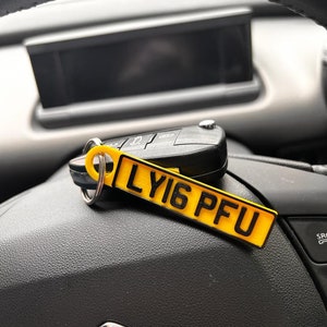 May include: A yellow car key chain with the text "LY16 PFU" printed on it. The keychain is hanging from a car key and is resting on a black leather surface.