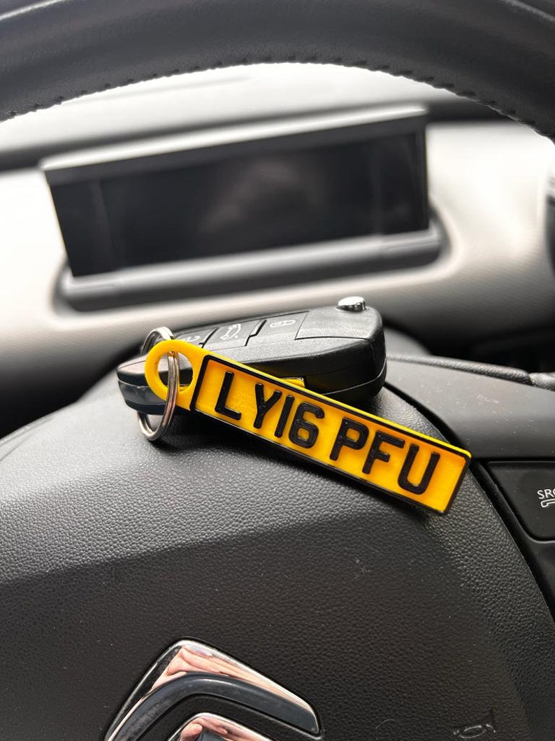 May include: A yellow car key chain with the text "LY16 PFU" printed on it. The keychain is hanging from a car key and is resting on a black leather surface.