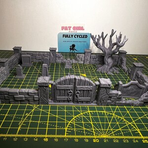 May include: A gray 3D printed miniature graveyard set with a gate, walls, tombstones, and a dead tree. The set is on a green cutting mat with a ruler.