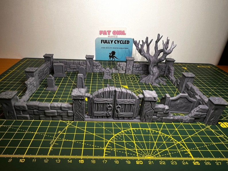 May include: A gray 3D printed miniature graveyard set with a gate, walls, tombstones, and a dead tree. The set is on a green cutting mat with a ruler.