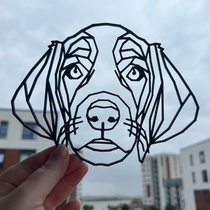 Op de afbeelding: Een zwart geometrisch honden gezicht ontwerp. Het gezicht van de hond is gemaakt met rechte lijnen, die een polygonale vorm vormen. De ogen, neus en oren van de hond zijn duidelijk gedefinieerd. De achtergrond is een bewolkte lucht en gebouwen.