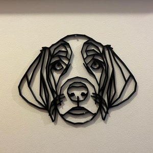 Op de afbeelding: Een zwart geometrisch metalen wandkunstwerk dat het gezicht van een hond afbeeldt. Het kunstwerk heeft een low-poly ontwerp, waarbij de kenmerken van de hond zijn gemaakt van verbonden lijnen. Het stuk is op een witte muur gemonteerd.