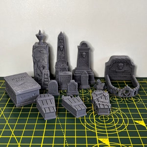 May include: A collection of 10 gray 3D printed miniature tombstones and coffins. The tombstones feature skulls, crosses, and the words "RIP" and "Here Lies". The coffins are made of wood and have a weathered look.