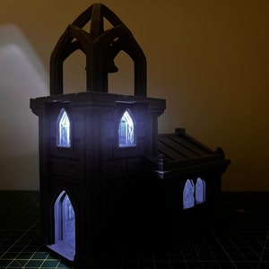 May include: A black 3D printed miniature church with a tower and windows. The windows are lit from inside, creating a warm glow.