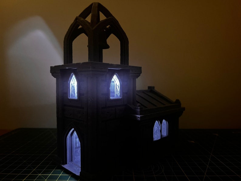 May include: A black 3D printed miniature church with a tower and windows. The windows are lit from inside, creating a warm glow.