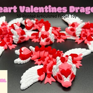 Puede incluir: Un juguete de fidget articulado impreso en 3D con forma de drag&oacute;n con alas rosas y blancas. El drag&oacute;n est&aacute; cubierto de peque&ntilde;os corazones rojos y tiene una cabeza en forma de coraz&oacute;n. El juguete se llama "Heart Valentines Dragon" y es un juguete de fidget articulado impreso en 3D.