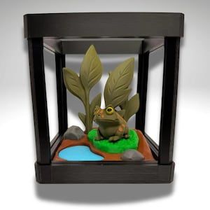 Puede incluir: Un terrario en miniatura con una figura de rana. La rana es verde con manchas naranjas y ojos amarillos, sentada sobre un parche verde. La escena incluye un estanque azul, rocas y plantas frondosas, todo dentro de un marco negro.