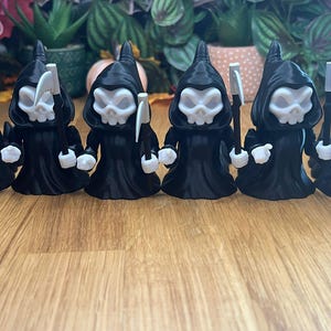 May include: Five black 3D printed figurines of Grim Reapers, each holding a scythe. The figurines are standing in a row on a wooden surface.