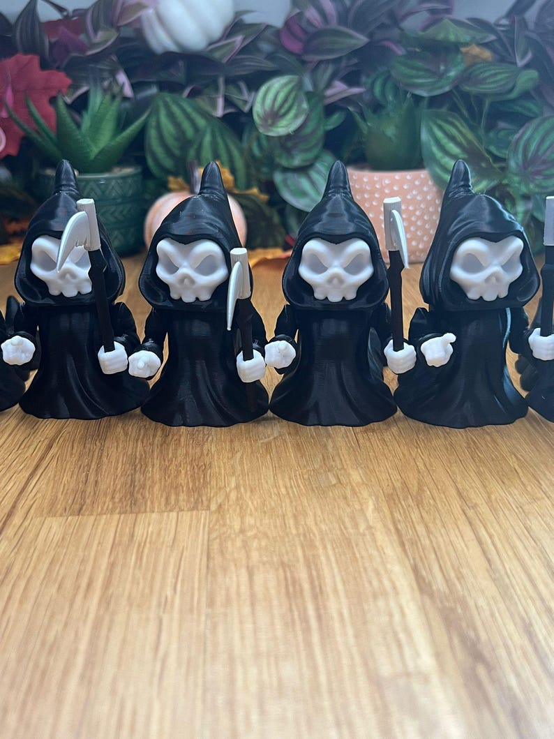 May include: Five black 3D printed figurines of Grim Reapers, each holding a scythe. The figurines are standing in a row on a wooden surface.