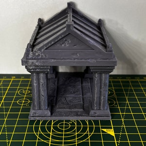 May include: A grey 3D printed miniature building with a gabled roof and columns. The building is sitting on a green cutting mat.