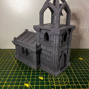 May include: A 3D printed gray miniature church with a bell tower and a pointed roof. The church has a small door and windows. The miniature is sitting on a green cutting mat.