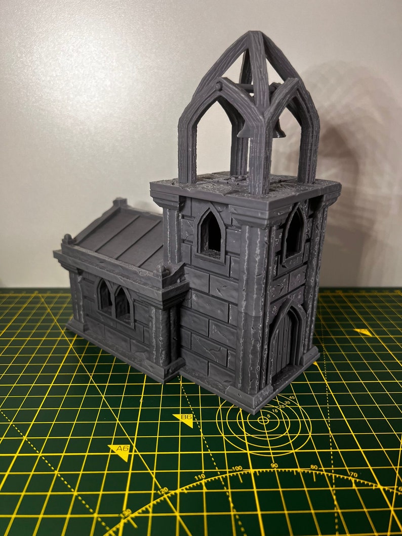 May include: A 3D printed gray miniature church with a bell tower and a pointed roof. The church has a small door and windows. The miniature is sitting on a green cutting mat.