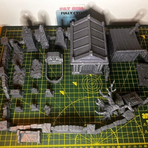 May include: A set of 3D printed miniature graveyard scenery pieces, including a mausoleum, a small building, a tree, and various tombstones and walls. The pieces are painted in a gray color and are arranged on a green cutting mat.