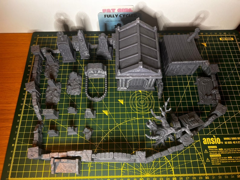 May include: A set of 3D printed miniature graveyard scenery pieces, including a mausoleum, a small building, a tree, and various tombstones and walls. The pieces are painted in a gray color and are arranged on a green cutting mat.