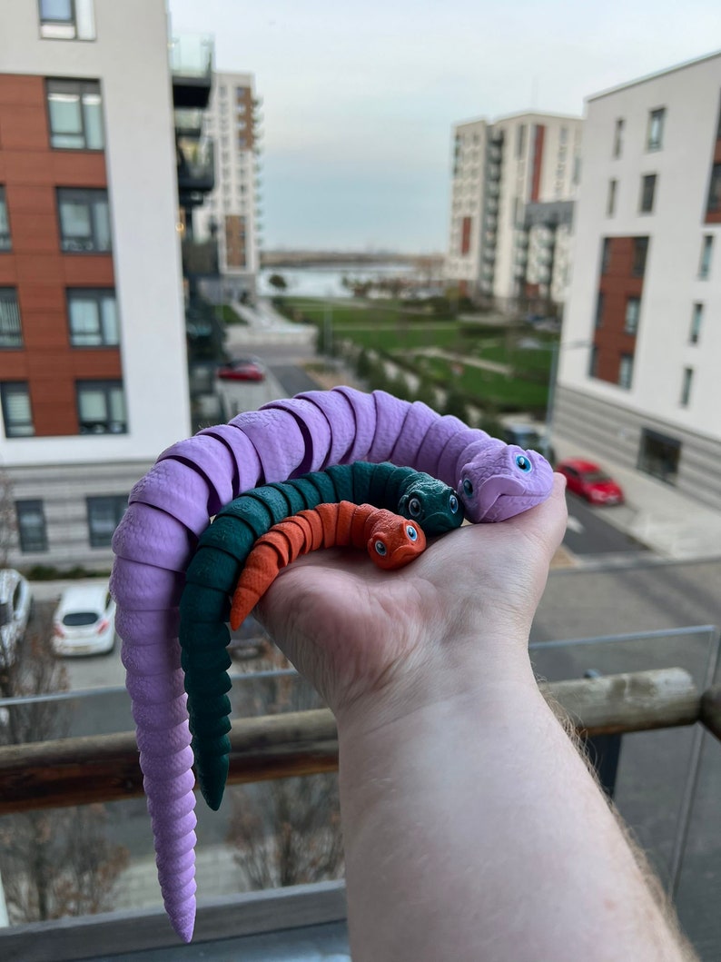 May include: Three colorful, segmented, bendable toys resembling worms. The largest worm is purple, the middle worm is green, and the smallest worm is orange. The worms are held in a person's hand.