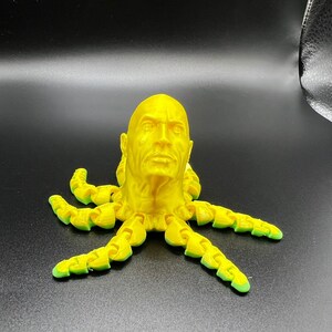 May include: A yellow 3D printed sculpture of a human head with a serious expression, attached to the top of an octopus with eight tentacles.