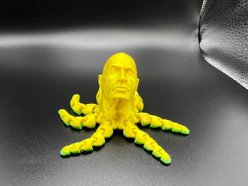 May include: A yellow 3D printed sculpture of a human head with a serious expression, attached to the top of an octopus with eight tentacles.