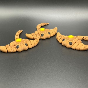 May include: Three brown and yellow 3D printed scorpion figurines. The scorpions have black eyes and are posed with their tails raised.