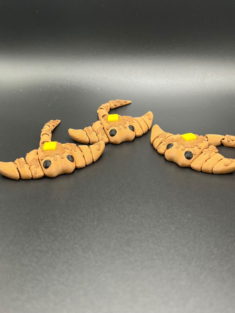 May include: Three brown and yellow 3D printed scorpion figurines. The scorpions have black eyes and are posed with their tails raised.