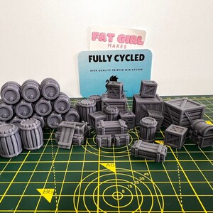 May include: A collection of gray 3D printed miniature barrels and crates. The barrels are stacked in a pyramid shape. The crates are of various sizes and shapes. The miniatures are on a green cutting mat. The text "FULLY CYCLED" is visible on a card in the background.