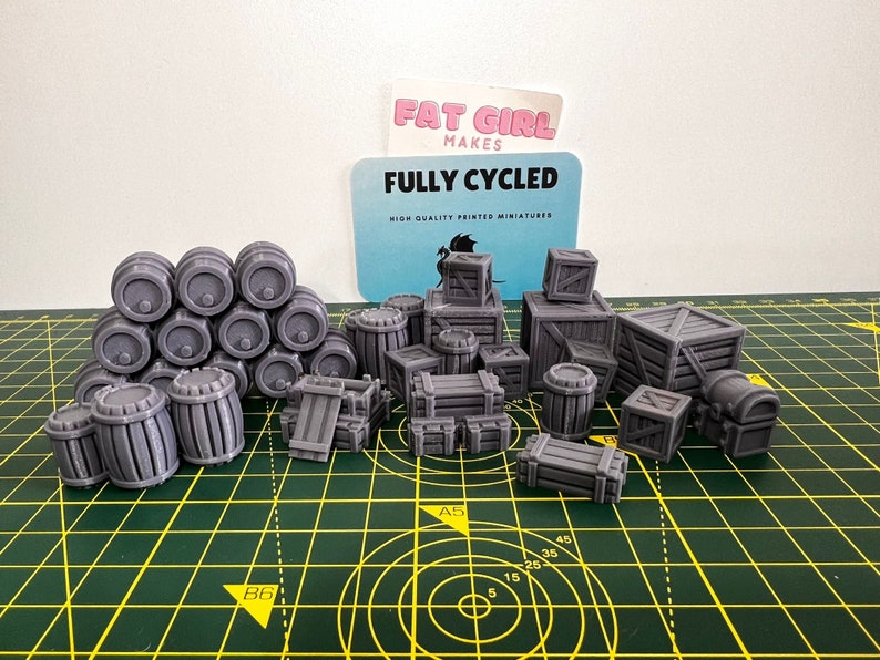 May include: A collection of gray 3D printed miniature barrels and crates. The barrels are stacked in a pyramid shape. The crates are of various sizes and shapes. The miniatures are on a green cutting mat. The text "FULLY CYCLED" is visible on a card in the background.