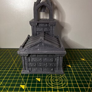 May include: A gray 3D printed miniature of a small church with a bell tower. The church has a pointed roof and a brick facade. The miniature is sitting on a green cutting mat.