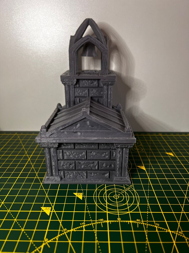 May include: A gray 3D printed miniature of a small church with a bell tower. The church has a pointed roof and a brick facade. The miniature is sitting on a green cutting mat.