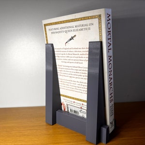 May include: A gray bookend with a book titled "Mortal Monarchs: Edge" standing on a wooden surface. The bookend is made of a sturdy material and has a simple design. The book is about the monarchs of England and Scotland.