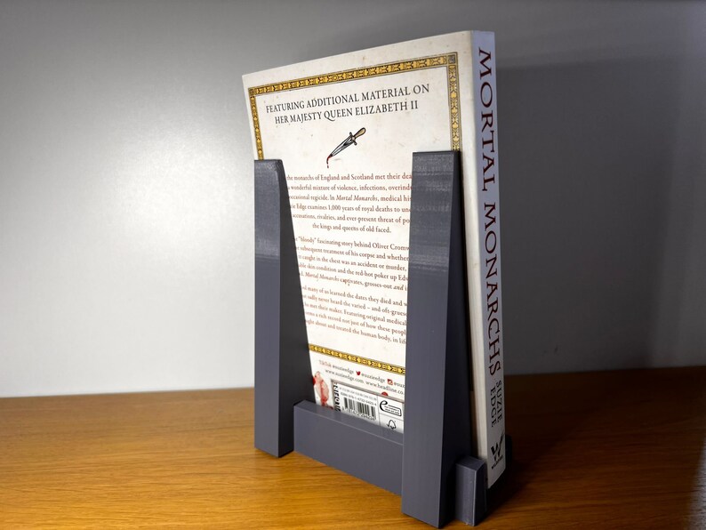 May include: A gray bookend with a book titled "Mortal Monarchs: Edge" standing on a wooden surface. The bookend is made of a sturdy material and has a simple design. The book is about the monarchs of England and Scotland.