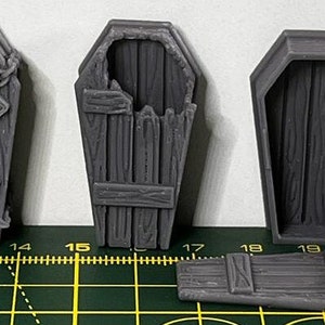 May include: Three grey wooden coffin-shaped boxes. One box has a chain and lock, another has a broken lid, and the third is closed. The boxes are on a green and yellow cutting mat.