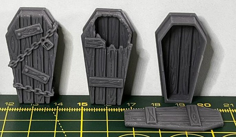 May include: Three grey wooden coffin-shaped boxes. One box has a chain and lock, another has a broken lid, and the third is closed. The boxes are on a green and yellow cutting mat.