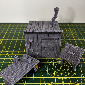 May include: A set of three gray 3D printed miniature buildings, including a small house with a chimney, a table with a phone, and a table with tools. The buildings are on a green cutting mat.