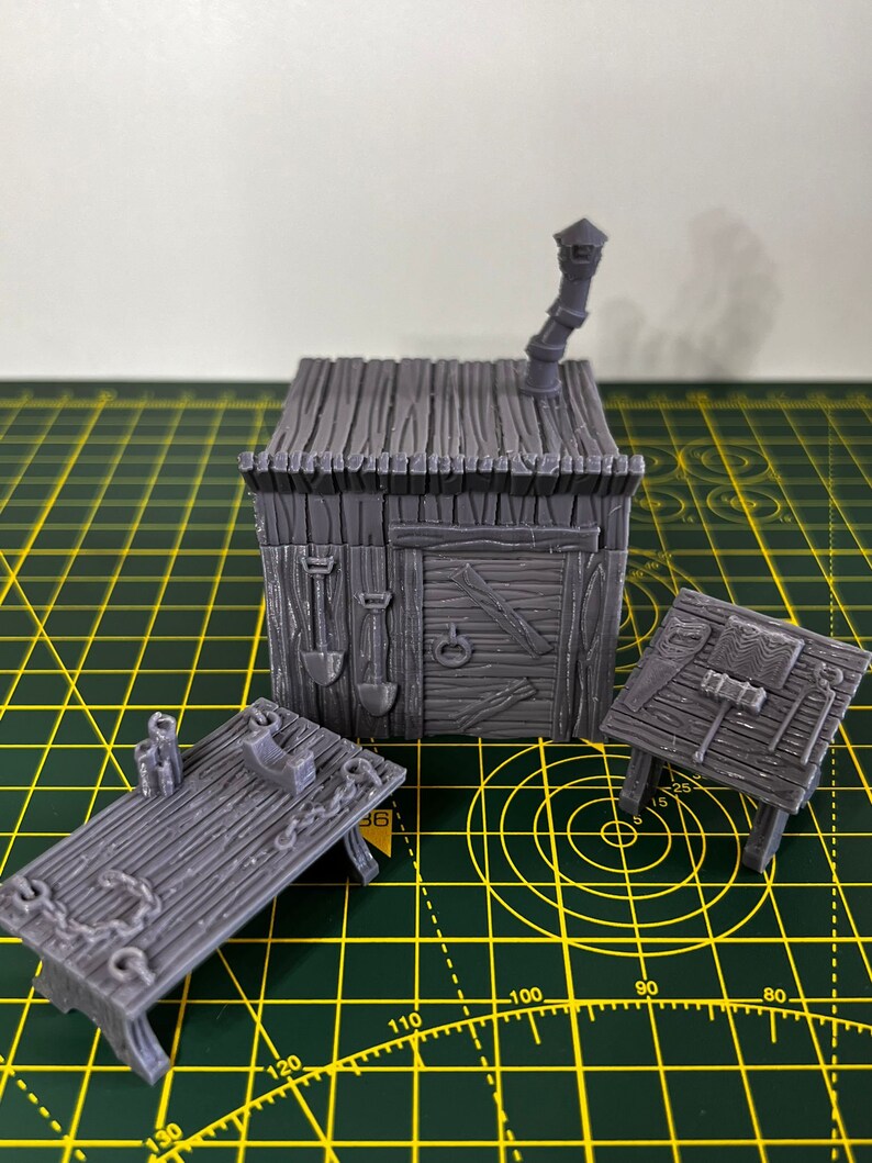 May include: A set of three gray 3D printed miniature buildings, including a small house with a chimney, a table with a phone, and a table with tools. The buildings are on a green cutting mat.