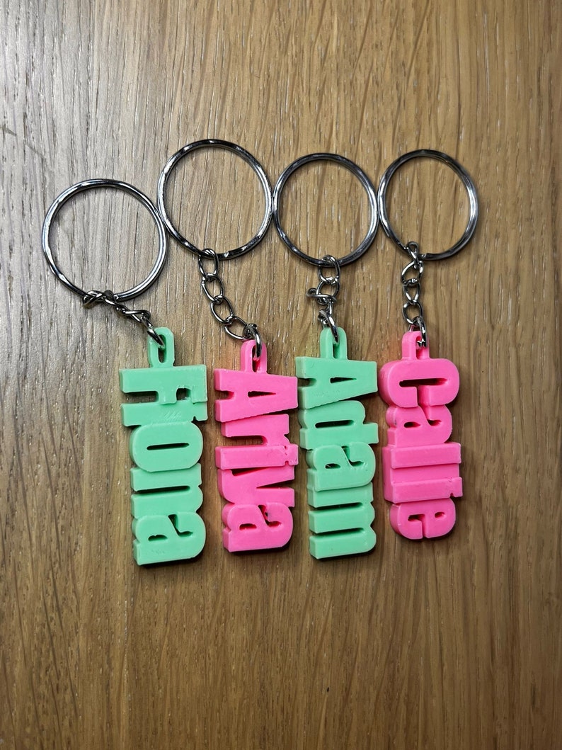 Op de afbeelding: Vier sleutelhangers met gepersonaliseerde namen. De namen zijn "Callie", "Austin", "Anna" en "Hope". De sleutelhangers zijn gemaakt van plastic en zijn roze en groen.