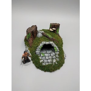 May include: A miniature stone tunnel entrance with a mossy green and brown exterior. The tunnel is surrounded by two tree stumps with green moss and mushrooms.