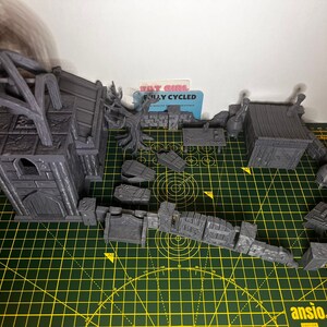 May include: A set of grey 3D printed miniature buildings for tabletop gaming. The set includes a church, a small house, and a variety of walls and other structures. The buildings are all made of a textured plastic and have a realistic look.