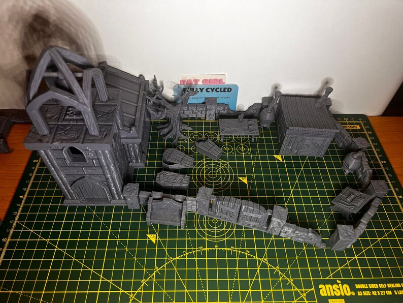 May include: A set of grey 3D printed miniature buildings for tabletop gaming. The set includes a church, a small house, and a variety of walls and other structures. The buildings are all made of a textured plastic and have a realistic look.