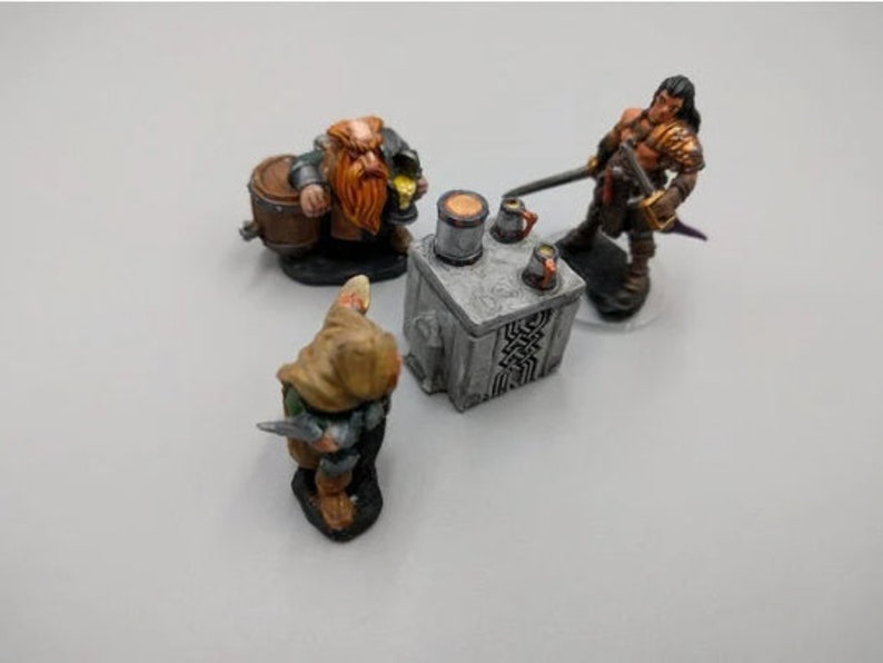 May include: A miniature scene with three figures: a red-haired dwarf holding a barrel, a cloaked figure with a dagger, and a warrior with a sword and shield. The figures are gathered around a small stone table with mugs and a tankard.